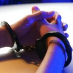 Close-up of handcuffed hands under blue police light, signifying arrest or detention.